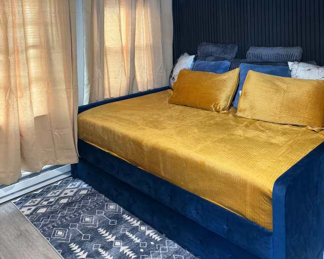 The image shows a cozy bedroom with a blue daybed covered in a mustard yellow bedspread, flanked by large cream curtains and featuring textured dark walls.