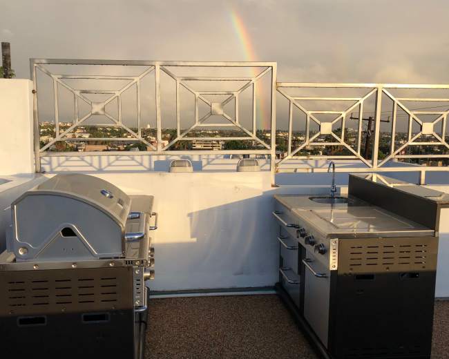 The image shows two stainless steel grills and a cooking station on a rooftop with a rainbow in the background.