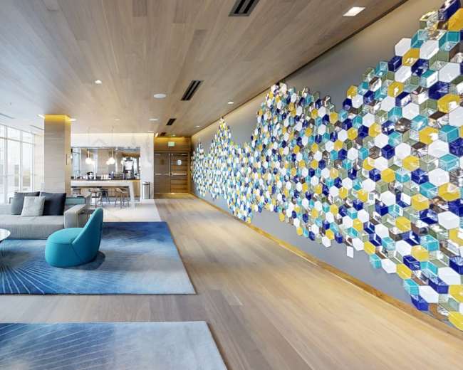 The image shows a modern interior space featuring a long corridor with a blue and yellow hexagonal tile wall, a seating area with a gray sofa and colorful chairs, and an open kitchen visible in the background.