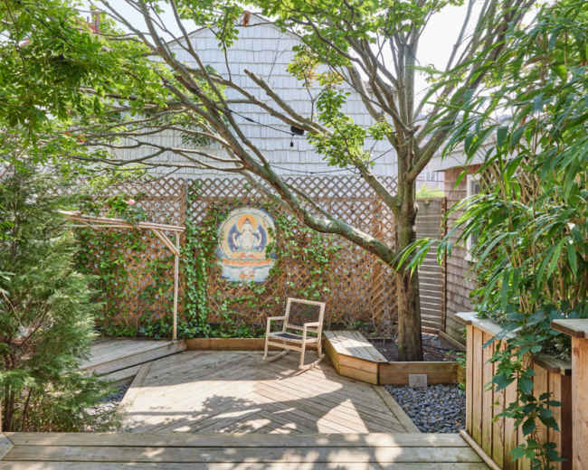 A wooden deck area surrounded by greenery features a single chair and a decorative mural on a lattice wall.