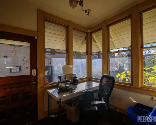 A desk with a chair sits in a sunlit room featuring large windows covered with blinds.