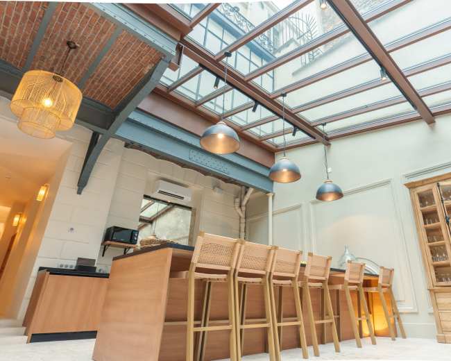 The image shows a modern kitchen with a glass ceiling, featuring wooden cabinets and a long dining table with high chairs.