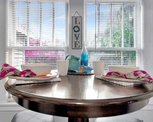 A wooden dining table is set for two with bowls, cups, and napkins, positioned near large windows adorned with blinds and overlooking outdoor plants.