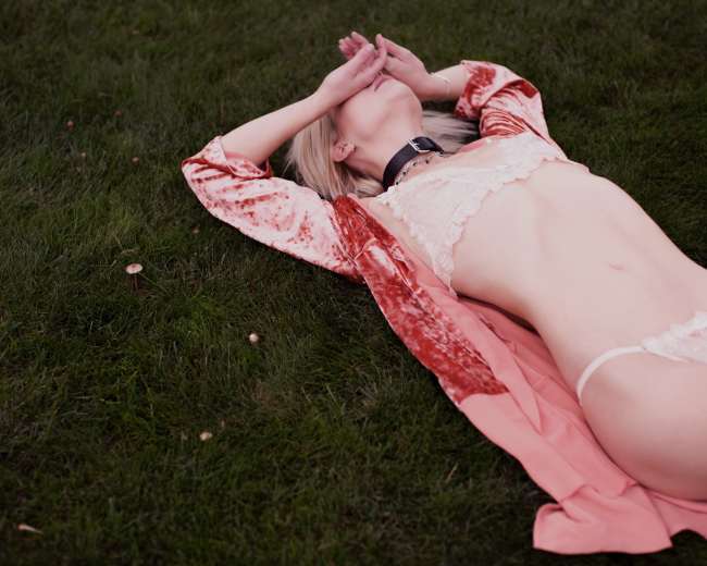 A person lies on grass wearing lingerie and a robe, with their hands covering their face.