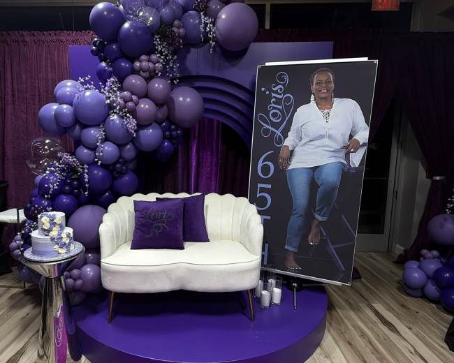 The image features a purple-themed seating area with a white couch, a large balloon arrangement, and a tall poster of a woman, all set against a purple backdrop.