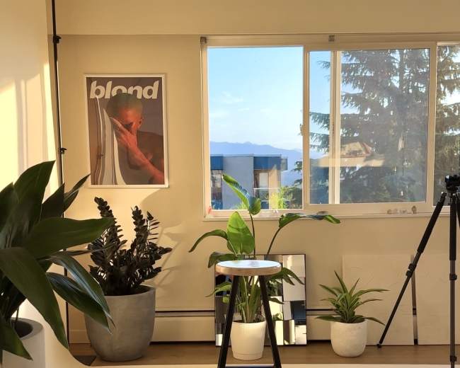 The scene depicts a bright and minimalistic studio space featuring potted plants, a stool, a camera tripod, and a large window with a view of distant hills.