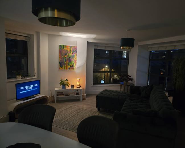 A modern living room features a sectional couch, a television on a white console, and large windows with city views.