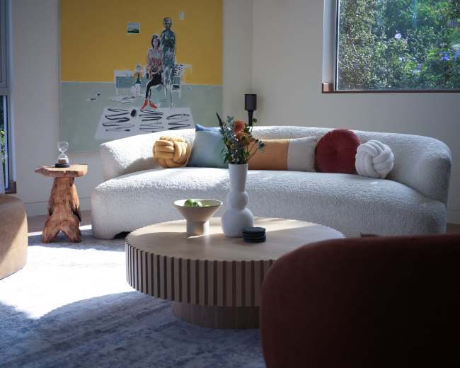The image shows a modern living room with a sculptural sofa, a round wooden coffee table, and a colorful painting on the wall.