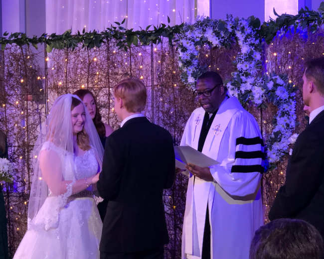 A couple exchanges vows during their wedding ceremony, surrounded by attendants and a officiant.