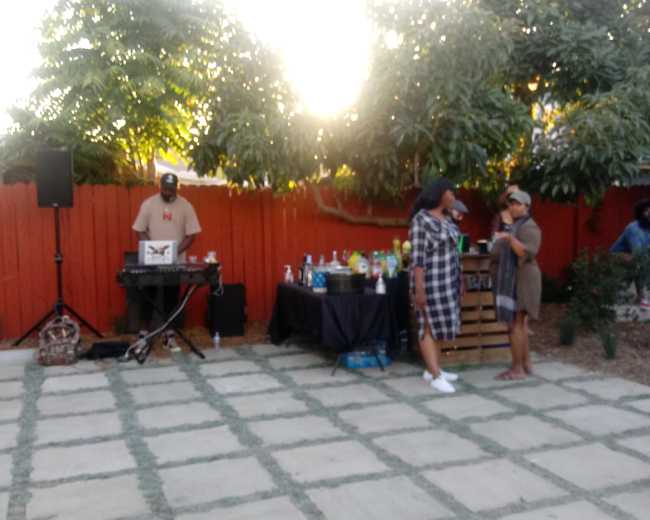 A backyard gathering features a DJ near a table of drinks, while guests chat and socialize.