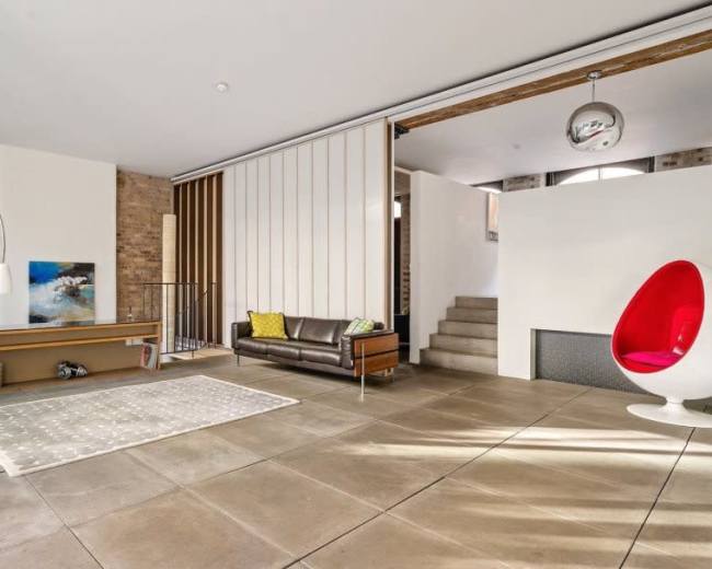 The image shows a modern living room with a brown leather sofa, a white rug, and a red pod chair, featuring exposed brick walls and a staircase leading to an upper level.