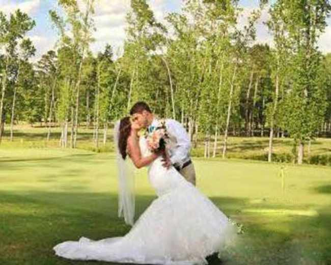 A bride and groom embrace in a romantic pose on a grassy field surrounded by trees.