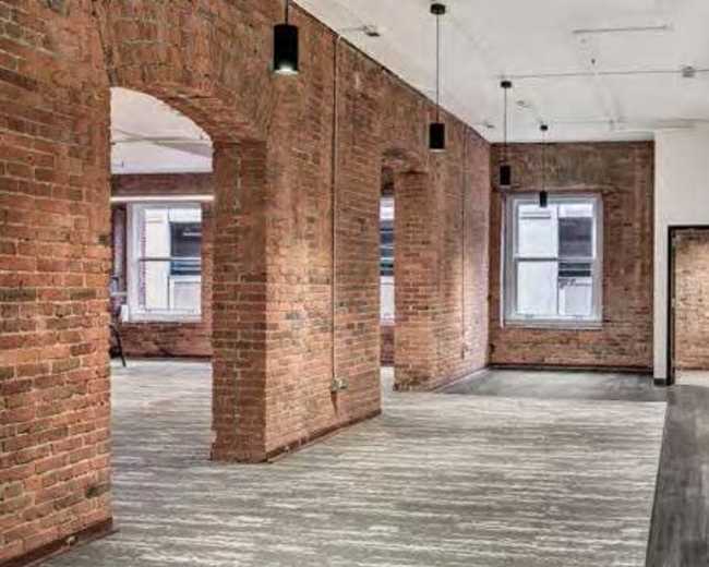 An empty commercial space with exposed brick walls and large windows, featuring gray flooring and modern light fixtures.
