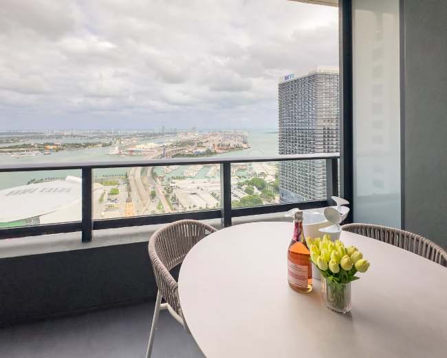 A balcony table set with a bottle of rose wine and a vase of yellow tulips overlooks a cityscape and waterfront.