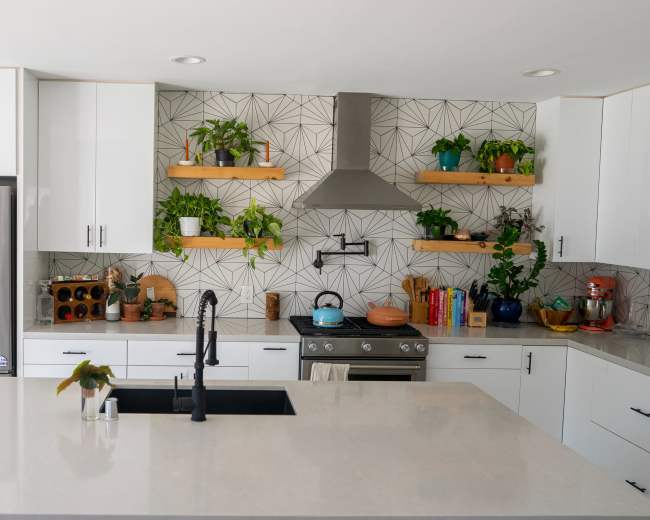 A modern kitchen with white cabinetry, a large island with a dark sink, and a patterned tile backsplash adorned with greenery on shelves.