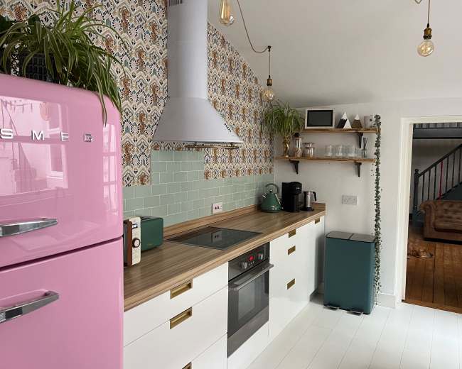The kitchen features a pink retro refrigerator, a modern stovetop, and patterned wallpaper, complemented by wooden countertops and green accents.