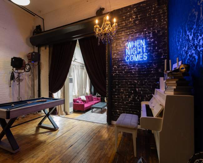 The image shows a room with a pool table, a neon sign that reads "WHEN NIGHT COMES," and a cozy seating area visible through a curtain.