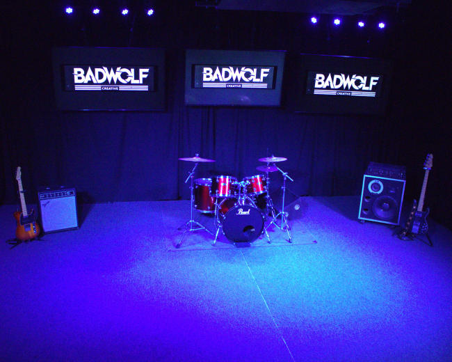 The image shows a music studio with a drum set, amplifiers, electric guitar on a stand, and three monitors displaying the logo "BADWOLF."