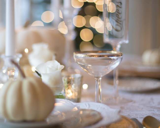 A elegantly set dining table features white pumpkins, roses, and sparkling glasses beside soft, blurred lights in the background.