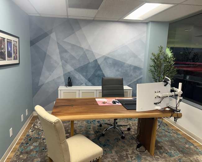 The image shows a modern office space featuring a wooden desk, ergonomic chair, computer monitor on an arm, and a decorative wall with geometric patterns.