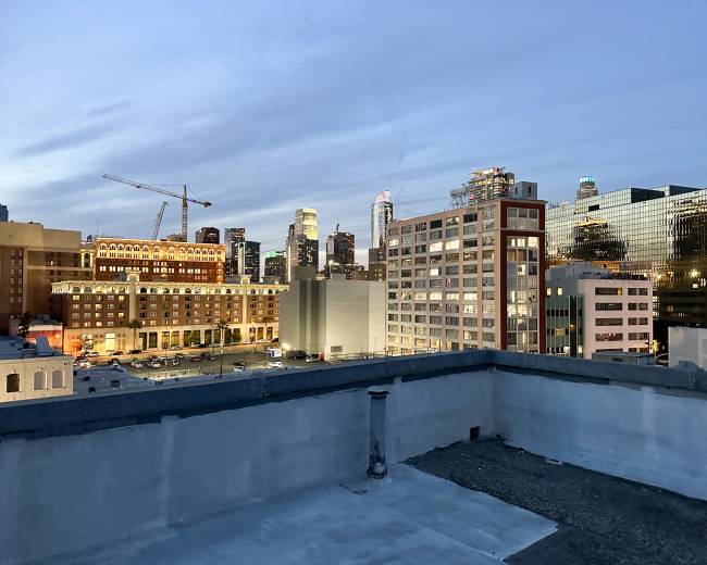 The image shows a rooftop view of a city skyline at dusk, with several buildings illuminated and construction cranes visible in the foreground.