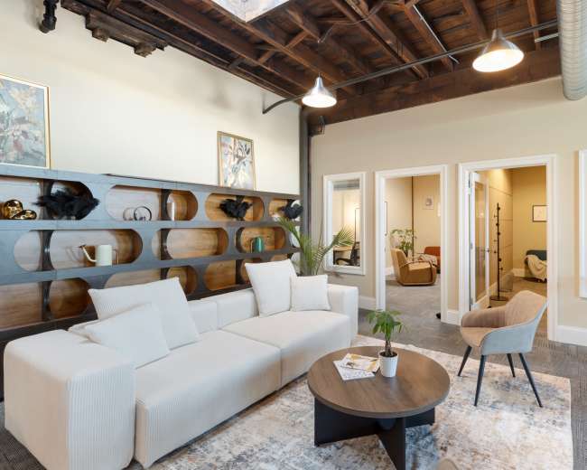 The image shows a spacious, modern living area featuring a white sectional sofa, a round coffee table, and a bookshelf with decorative items, all under a ceiling with exposed wooden beams.