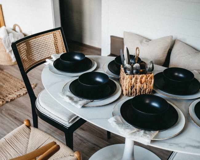 A round dining table is set with black bowls and plates, accompanied by a basket of utensils, in a softly lit room.