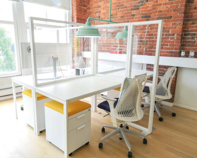 A bright office space with two workstations divided by clear plastic partitions and equipped with ergonomic chairs and storage units.