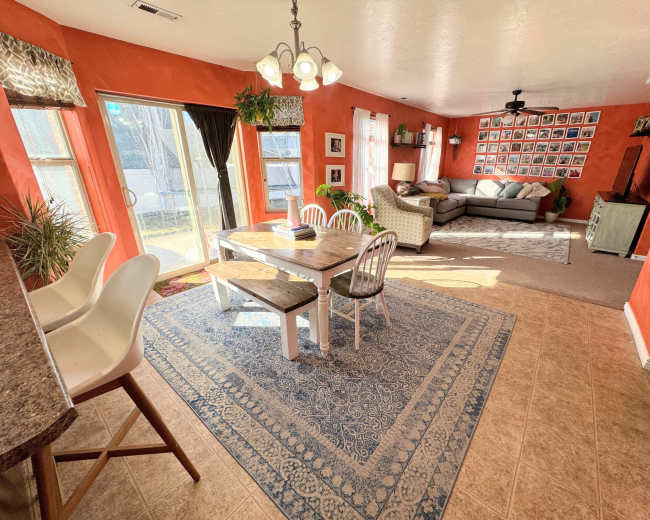 The image shows a brightly colored room with a dining table, chairs, and a living area featuring a sofa and numerous framed photos on the wall.