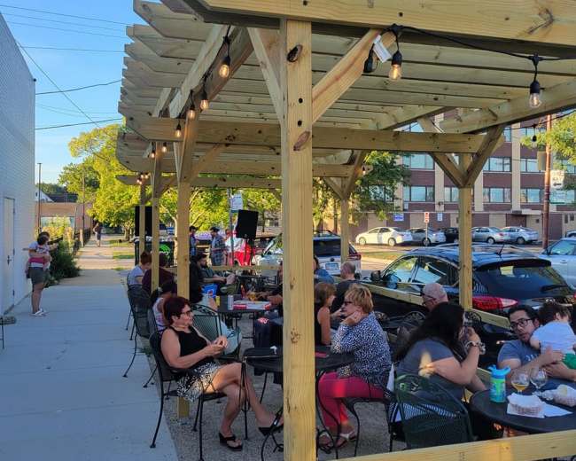 People are gathered at outdoor tables under a wooden pergola with string lights, enjoying a warm evening in a parking lot setting.