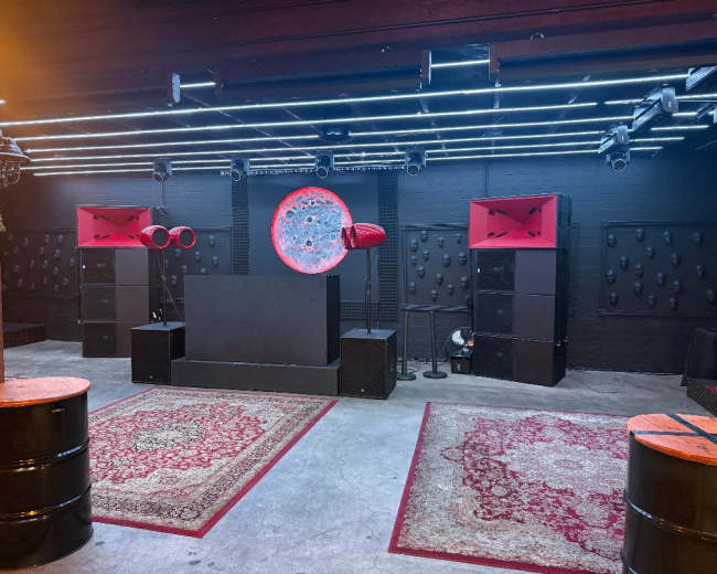The image shows a music venue setup with a stage, large speakers, and patterned rugs on a concrete floor.