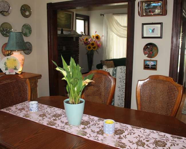 A wooden dining table with a pink table runner and a potted plant is set in a room adorned with art on the walls and a view into a living area.