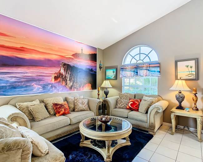 A living room features a large wall mural of a coastal sunset, with beige sofas, a glass coffee table, and lamps beside the windows.