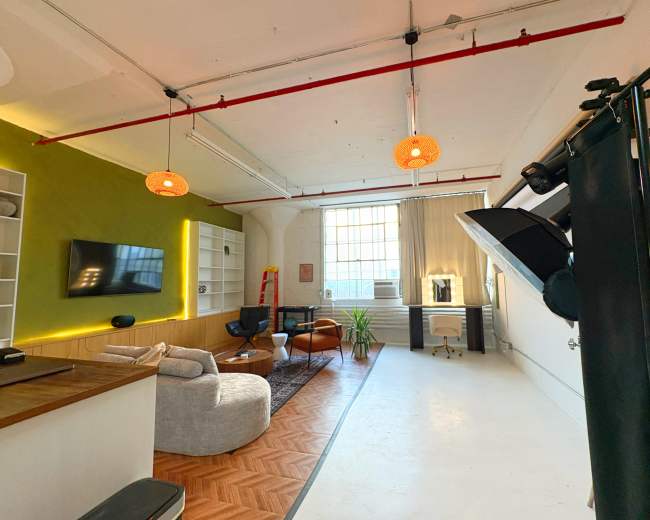 The image shows a well-lit studio apartment with a green accent wall, a seating area, a large window, and minimalistic decor.