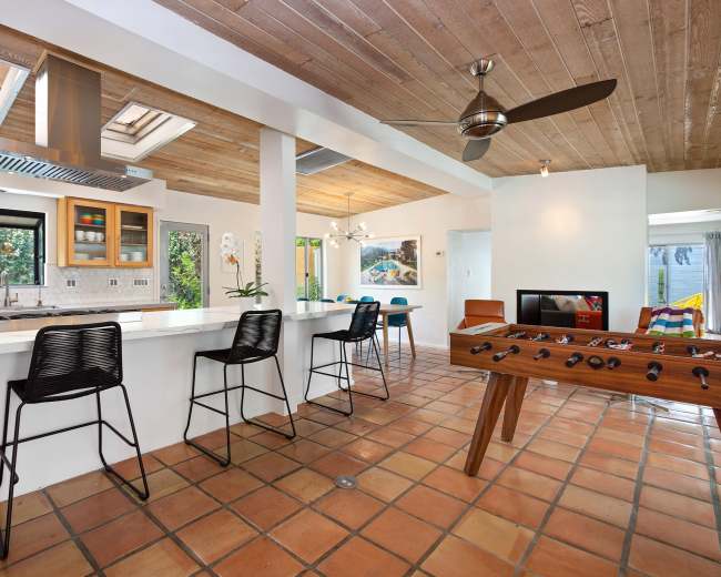 The image shows a modern kitchen and living area with a tiled floor, bar seating, and a foosball table.