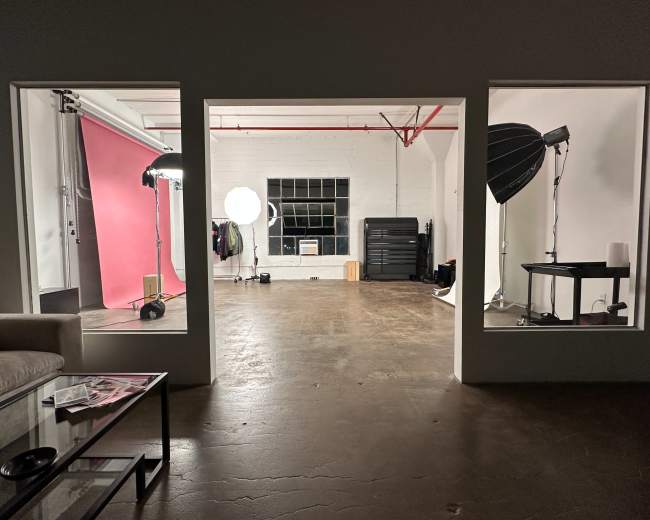 The image shows a spacious studio with three open windows, featuring a pink backdrop, lighting equipment, and polished concrete floors.