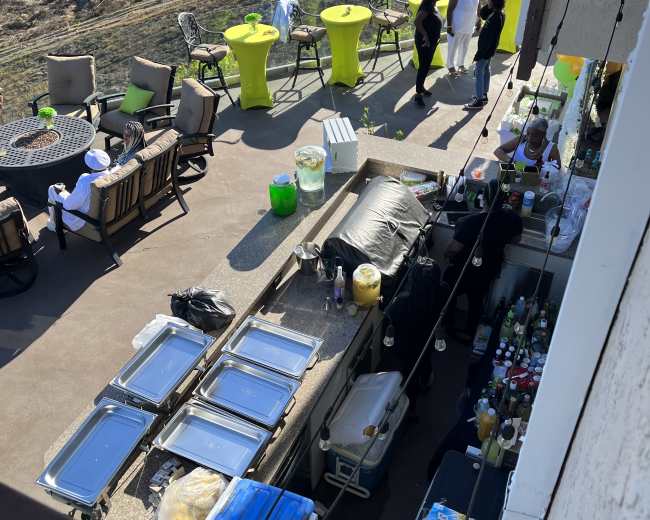 The image shows an outdoor event setup with tables and chairs, featuring a bar area stocked with various drinks and food containers.