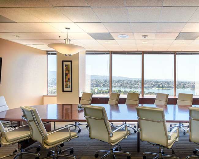 A large conference room features a long table surrounded by white chairs, with large windows providing a view of the city and mountains in the distance.