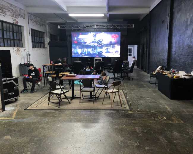 A large screen displays images in an industrial-style room with chairs and tables arranged around a carpet.