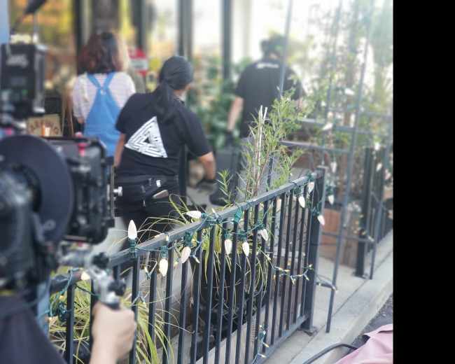 A film crew sets up equipment while two individuals walk past a railing adorned with plants at a filming location.
