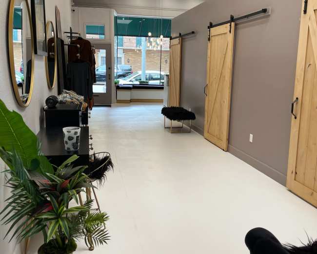 The image shows a narrow, well-lit hallway of a retail space with wooden sliding doors, a decorative plant in a pot, and minimalist furniture along the walls.