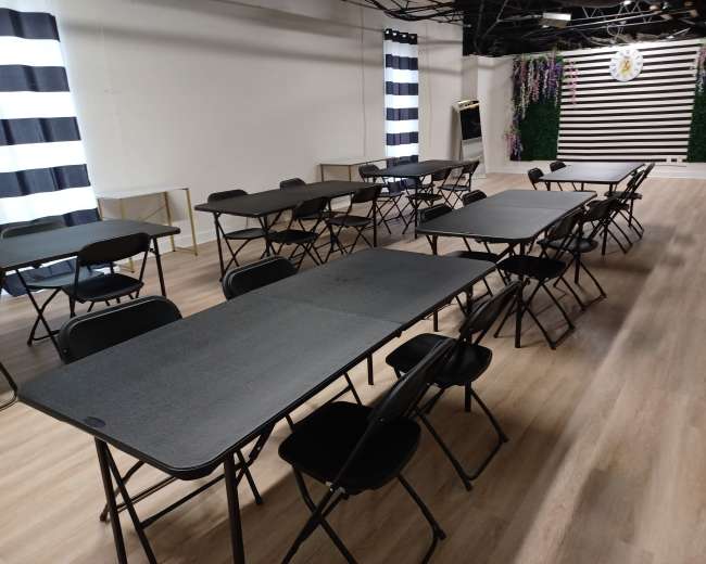 The image shows a room with several black folding tables and chairs arranged in rows, with striped curtains in the background.
