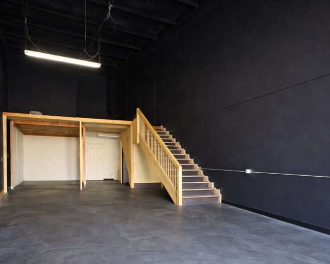The image shows an empty interior space with dark walls, a wooden staircase on the right, and a small area with a door and platform in the back.