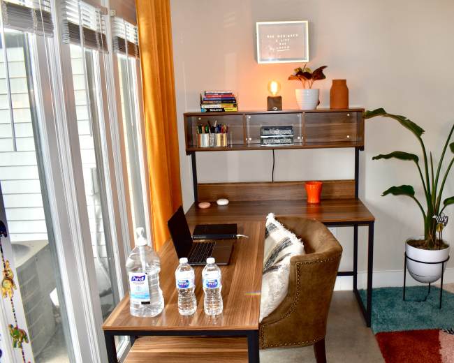 The image shows a home office setup featuring a wooden desk with a laptop, three water bottles, and a decorative plant beside it, set against a light-colored wall with large windows.