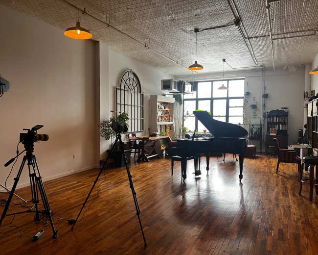 A spacious room featuring a grand piano, camera equipment on tripods, and various plants, with ample natural light coming through large windows.