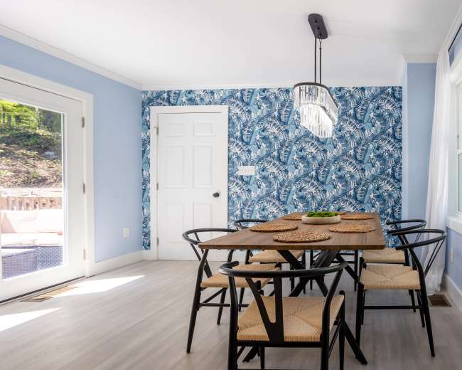 A dining area features a wooden table surrounded by six black chairs, with a blue floral wallpaper and a sliding glass door leading outside.