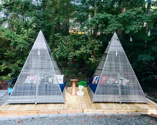 The image shows two triangular metal structures with open sides, placed on a wooden deck surrounded by trees.