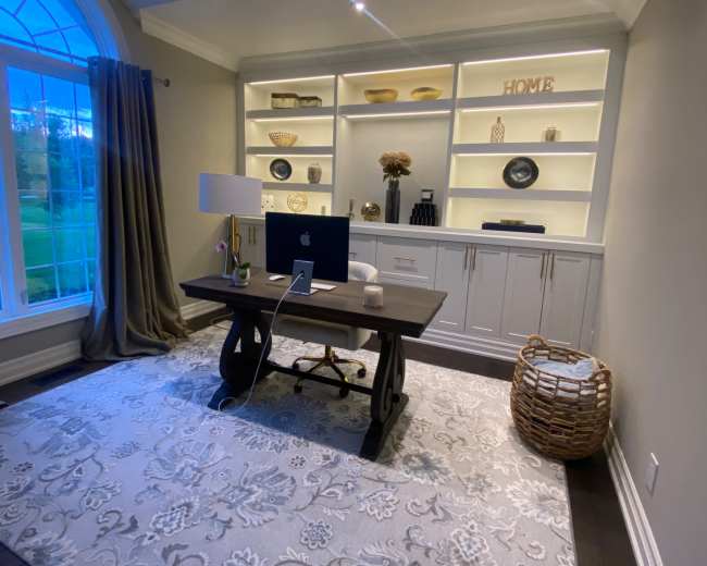 The image shows a neatly arranged home office featuring a desk with a computer and a lamp, surrounded by built-in shelving and a decorative area rug.