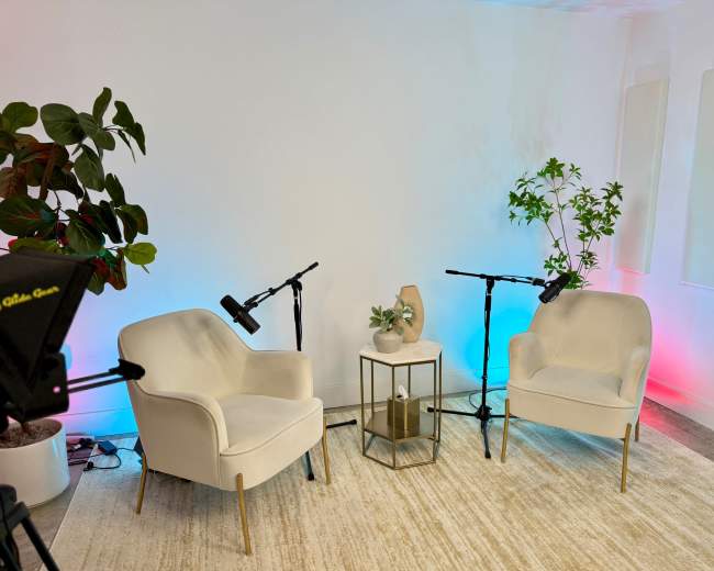 The image shows a minimalistic interview or discussion setup with two white armchairs, a small table, and two microphones on stands, surrounded by green plants and softly lit walls.