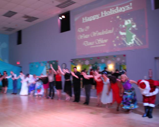 A group of dancers in formal attire performs in a spacious hall decorated for a holiday event, with a large screen displaying "Happy Holidays!" in the background.